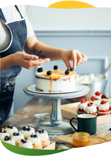 A person, standing in the kitchen, is decorating a cake with berries. On the work surface are several cupcakes