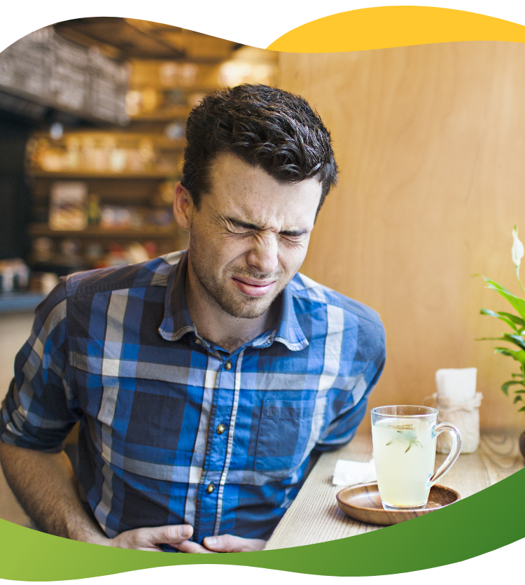 A young man sits at the window seat in a cafe and grips his belly with a pained face - his drink in front of him is empty