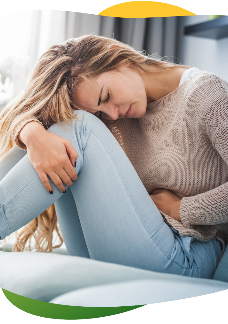 A young woman suffering from period bloating and sitting cramped on the floor holding her abdomen with her left hand